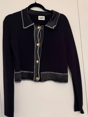 Svart kofta från Claudie Pierlot - Stilren svart kofta från Claudie Pierlot med vita kontrastsömmar och dekorativa guldfärgade knappar. Koftan har lång ärm, krage och ett elegant mönster längs kanterna. Perfekt för dig som gillar klassisk men trendig stil.