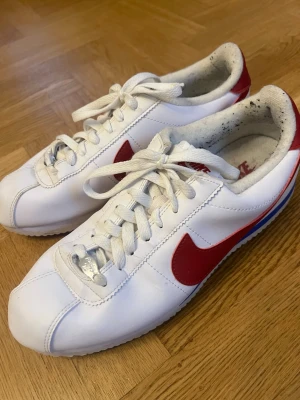 Nike Cortez vita och röda sneakers - Klassiska Nike Cortez sneakers i vitt läder med röda detaljer och blå rand på sulan. Skorna har lågt skaft, vita skosnören och den ikoniska Nike-loggan på sidan och hälen. Perfekta för en sportig och retro look. Se skick på bild.
