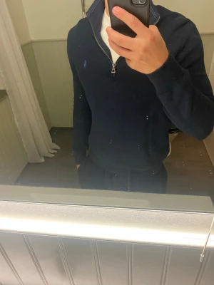 Mörkblå halvzip tröja från Polo Ralph Lauren - Snygg mörkblå stickad tröja från Polo Ralph Lauren med halv dragkedja och klassisk broderad logga på bröstet. Tröjan har hög krage och ribbade muddar vid ärmslut och nederkant. Perfekt för en stilren och avslappnad look.