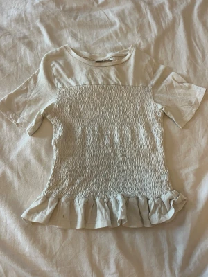 Vit topp med volang från Gina Tricot - Supersöt vit topp från Gina Tricot med smockad framsida och volangkant nedtill.
