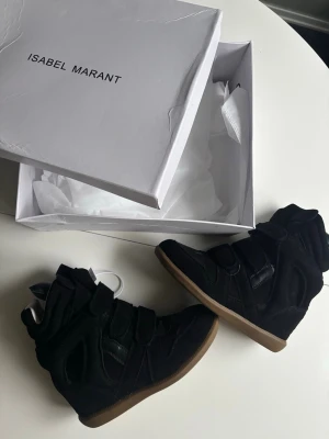 Svarta Isabel Marant sneakers highs - Snygga svarta sneakers från Isabel Marant med högt skaft och cool design. Skorna har en mix av mocka och läderdetaljer samt kardborreband över foten. Yttersulan är i gummi med en klassisk brun färg. Perfekta för dig som vill ha en statement sneaker.