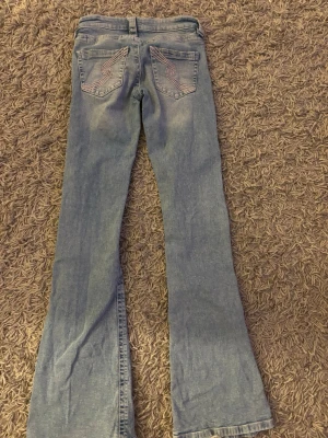 Ljusblå bootcut jeans med broderi - Säljer ett par ljusblå bootcut jeans med snyggt broderi på bakfickorna. Jeansen har klassisk femficksmodell och är tillverkade i mjukt denimtyg. Perfekta för dig som gillar retrovibbar och vill ha ett par jeans med unik detalj på fickorna.