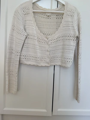Vit kort stickad cardigan - Söt vit stickad cardigan med lång ärm och knappar framtill. Tröjan har ett hålmönster i ränder och vågig kant längs halsringning och nederkant. Croppad modell som passar perfekt till högmidjade jeans eller kjol. Materialet känns mjukt och luftigt.