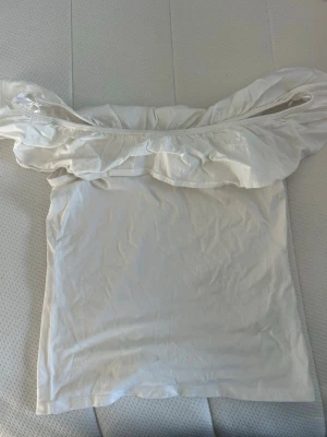 Vit offshoulder topp med volang - Säljer en vit axelbandslös topp med bred volangkant upptill. Toppen är offshoulder och har en figurnära passform. Perfekt för dig som gillar en enkel men ändå söt stil.