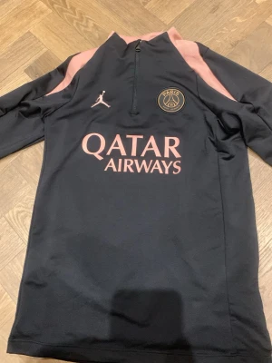 Svart PSG träningströja Jordan - Snygg svart träningströja från PSG x Jordan med rosa detaljer och halv dragkedja. Tröjan har Paris Saint-Germain-logga på bröstet och Qatar Airways-tryck framtill. Tillverkad i Dri-FIT-material som andas, perfekt för träning eller sport.