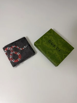 Svart plånbok med orm - Säljer en svart plånbok från  med GG-mönster och en cool röd, vit och svart orm på framsidan. Plånboken är fyrkantig och tillverkad i slitstarkt material, perfekt för dig som vill ha något unikt och trendigt. Kommer med original grön box. 