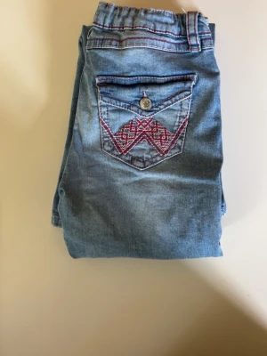 Ljusblå jeans med broderad ficka - Säljer ett par ljusblå jeans med snygga slitningar och broderad bakficka i rosa och rött mönster. Jeansen har resår i midjan och kontrastsömmar i rosa. Perfekta för en avslappnad och trendig look.