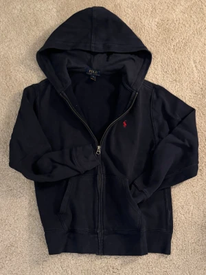 Mörkblå hoodie från Polo Ralph Lauren - Snygg mörkblå hoodie med dragkedja från Polo Ralph Lauren. Klassisk design med huva, fickor framtill och den ikoniska röda logotypen broderad på bröstet. Tillverkad i mjukt bomullsmaterial som är skönt att ha på sig. Perfekt för en avslappnad stil. (Ser svart ut på bilderna men är mörkblå)