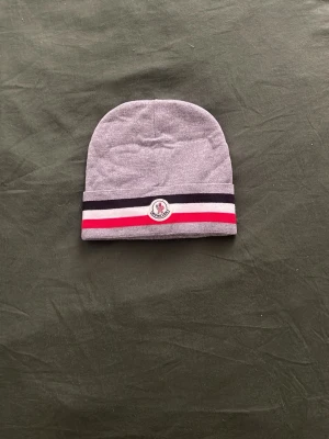 Grå Moncler mössa  - Snygg grå mössa från Moncler med svart, vit och röd rand runt kanten samt Moncler-logga framtill. Klassisk modell i mjukt material som passar perfekt till kalla dagar. Enkel att matcha med olika outfits.