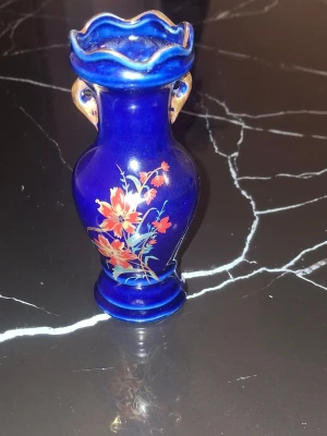 Blå vas med blommotiv i glas - Snygg blå glasvas med handtag och vågig kant upptill. Vasen har ett handmålat blommotiv i rött, orange och vitt på framsidan. Perfekt som dekoration eller för mindre buketter. Materialet är glas och färgen är djupblå med detaljer i guld.