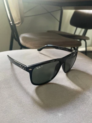 Svarta Ray-Ban boyfriend - Snygga svarta solglasögon från Ray-Ban med klassisk wayfarer-form och mörka glas. Bågarna är i blank svart plast och har Ray-Ban-loggan på sidan samt i hörnet av linsen. Perfekta för att höja din stil i solen. Nya!