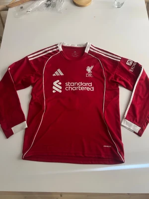 Liverpool 25/26 Szoboszlai  - Liverpool FC långärmad matchtröja i rött med vita detaljer och tryck. Tröjan har Adidas-logga, klubbmärke och sponsortryck fram samt namn och nummer 'Szoboszlai 8' på ryggen. Tillverkad i funktionsmaterial med Aeroready-teknologi och klassiska ränder på ärmarna.