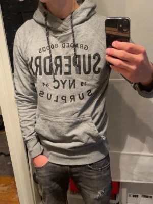 Grå hoodie från Superdry - Snygg grå hoodie från Superdry med svart tryck framtill med texten ”superdry NYC Surplus. Perfekt för dig som vill ha en lite mer archive stil. Modellen är 170cm och väger 64 klio.