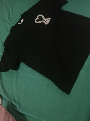 Ami Paris T-shirt  - Säljer en svart t-shirt med en vit hjärtdetalj på bröstet. T-shirten har klassisk passform och korta ärmar. Perfekt till jeans eller shorts för en enkel och stilren look.