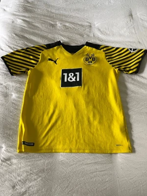 Gul Borussia Dortmund matchtröja Puma Bellingham  - Snygg gul Borussia Dortmund matchtröja från Puma med svarta detaljer och ränder på ärmarna. Tröjan har klubbmärke och sponsorlogga framtill samt korta ärmar. Perfekt för fotbollsträning eller att bära till vardags.