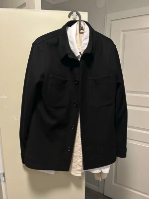 Svart overshirt med fickor - Stilren svart overshirt med klassisk krage och två stora bröstfickor. Jackan har raka ärmar och knäpps med svarta knappar framtill. Perfekt lager-på-lager-plagg för en trendig look. Materialet ser ut att vara kraftigt tyg, vilket ger en snygg och strukturerad passform.