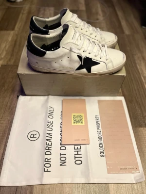 Golden Goose - Golden Goose sneakers i vitt läder med marinblå stjärndetalj på sidan och marinblå hälparti med logga. Klassisk låg modell med slitna detaljer för en trendig look. Skorna har vita snören och perforerade hål på sidan för ventilation.