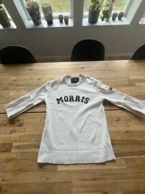 Vit stickad tröja från Morris - Vit stickad tröja från Morris med svart text på bröstet. Tröjan har rund halsringning, ribbade muddar och bred ribbad nederkant. Ärmarna är trekvarts och har diskreta detaljer vid armbågen. Perfekt för en clean och stilren look.