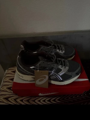 Grå Asics sneakers med mesh och silverdetaljer - Säljer ett par grå Asics sneakers med mesh-ovandel och silverfärgade detaljer. Skorna har vita sulor och klassisk snörning. Perfekta för dig som gillar sportig stil och bekväma skor. Taggen sitter kvar på skon.
