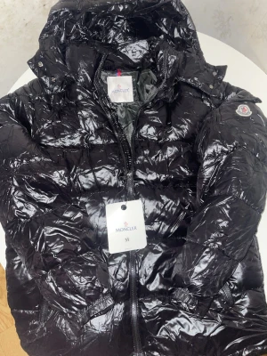 Svart glansig dunjacka från Moncler - Säljer en svart glansig dunjacka från Moncler med huva och dragkedja framtill. Jackan har Moncler-logga på ärmen, fickor med lock och knapp samt quiltad design. Perfekt för kalla vinterdagar och riktigt snygg streetstil.