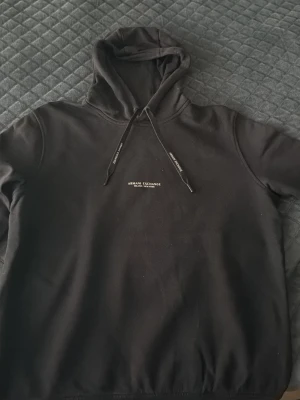 Svart hoodie från Armani Exchange - Snygg svart hoodie från Armani Exchange med justerbar huva och dragsnören med logga. Diskret tryck på bröstet med texten 'Armani Exchange Milano/New York'. Perfekt för en clean och stilren look.