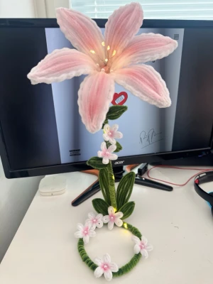 Handgjord blomlampa med LED - Unik handgjord blomlampa med stor rosa och vit blomma i tyg, gröna blad och små dekorationsblommor på stjälken. Lampan har inbyggda LED-ljus som lyser upp blomman och ger ett mysigt sken. Perfekt som dekoration på skrivbord eller hylla. Drivs med batteri.