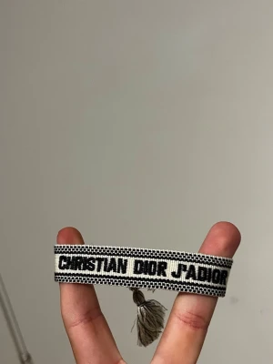 Christian Dior J'Adior tygarmband - Snyggt tygarmband från Christian Dior med texten 'J'Adior' i svart på beige botten. Armbandet har en vävd design och avslutas med tofsar i matchande färger. Perfekt accessoar för att lyfta din outfit med en exklusiv touch.