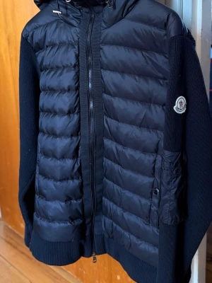 Svart pufferjacka med huva Moncler - Snygg svart pufferjacka från Moncler med quiltad framsida, stickade ärmar och rygg samt en praktisk huva. Jackan har dragkedja framtill, sidofickor och Moncler-logga på ärmen. Perfekt för kalla vinterdagar och riktigt stilren look.