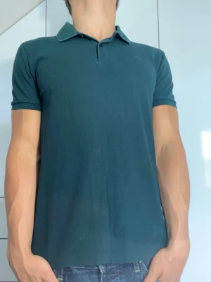 Mörkgrön piké från J.Lindberg - Mörkgrön piké från J.Lindberg i 100% bomull. Strl S. Modellen är 178 cm och väger 77 kg. Hör gärna av dig vid frågor!