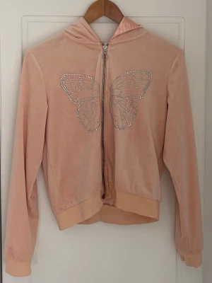 Ljusrosa velour hoodie med fjäril strl 170 - Säljer en ljusrosa hoodie i mjuk velour med dragkedja och huva. På framsidan finns en stor fjäril i glittrande strasstenar som ger en extra snygg detalj. Hoodien har ribbade muddar och en avslappnad passform. Perfekt för dig som gillar Y2K-stil!