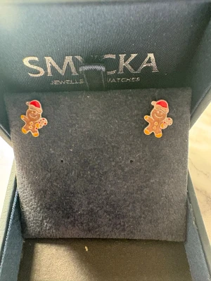 Pepparkaksgubbe örhängen med tomteluva - Säljer ett par söta örhängen formade som pepparkaksgubbar med röda tomteluvor och vita detaljer. Perfekta för dig som vill ha en lekfull och julig touch på din stil. Örhängena är små och färgglada, tillverkade i metall med emalj.