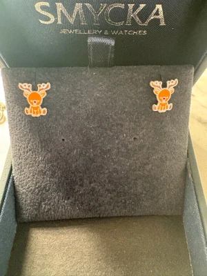 Söta renörhängen i orange och vitt - Unika örhängen formade som små renar i orange och vitt. De har en lekfull design med detaljerade horn och ansikte, perfekta för dig som vill ha något gulligt och annorlunda i smyckeskrinet.
