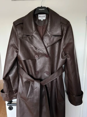 Brun trenchcoat i skinnimitation NA-KD - Snygg brun trenchcoat från NA-KD i skinnimitation. Ser exakt ut som ny, enbart använd 1-2 gånger.  Nypriset var 1500kr. Lång modell med dubbelknäppning, bred krage och avtagbart midjebälte. Jackan har två stora fickor framtill och justerbara remmar vid ärmsluten för extra stilpoäng. Otroligt snygg!