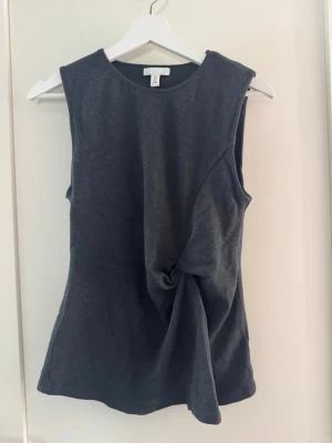 Mörkgrå ärmlös topp med knyt - Snygg mörkgrå ärmlös topp med rund halsringning och unik knytdetalj på sidan. Toppen har en clean look och är tillverkad i ett mjukt material som känns skönt mot huden. Perfekt för dig som gillar minimalistisk stil med en twist. Storlek S från H&M. Använd en gång, därav nyskick!