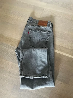 Grå raka jeans med läderpatch - Säljer ett par grå jeans med rak passform och klassisk femficksdesign. Jeansen har en brun läderpatch bak i midjan och en röd detalj vid bakfickan. Materialet är denim i bomull och färgen är tvättad grå, perfekt till en avslappnad stil.