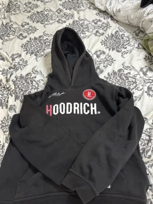 Svart hoodie från Hoodrich - Säljer en svart hoodie från Hoodrich med stor logga i vitt och rosa på bröstet samt en röd rund patch med H. Klassisk huva och magficka framtill. Hoodien har ribbade muddar vid ärmslut och nederkant. Perfekt för en streetwear-look.   pris kan diskuteras 