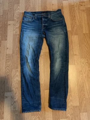 Nudie jeans grim Tim  - Snygga mörkblå jeans med tydliga slitningar och ljusare partier på låren och knäna. Tydlig defekt på bakficka. Sömmen på undersidan syns ej när man har på sig jeansen. Storlek W31 L34