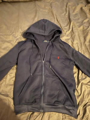 Mörkblå hoodie från Polo Ralph Lauren - Mörkblå hoodie med dragkedja från Polo Ralph Lauren. Klassisk design med huva och dragsnören, två fickor framtill och den ikoniska röda logotypen broderad på bröstet. Perfekt för en avslappnad och stilren look.
