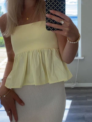 Ljusgul axelbandslös peplum tubtopp - Söt ljusgul axelbandslös tubtopp med peplumvolang. Överdelen är ribbad och stretchig medan den nedre delen har ett glansigt, lätt tyg som ger en luftig känsla. Perfekt för varma dagar och enkel att styla med kjol eller jeans.