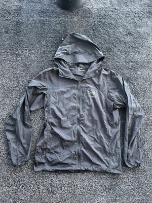 Arc'teryx vindjacka - Snygg grå vindjacka från Arc'teryx med huva och dragkedja framtill. Jackan har två sidofickor med dragkedja och en lätt, tunn design som passar perfekt för blåsiga dagar. Logotyp på bröstet och justerbar huva för extra skydd. Storlek M, vet inte mycket om den, skick 9/10. Pris går att diskutera✅