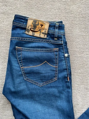 Jacob Cohen jeans  - Snygga blå jeans från Jacob Cohen med klassisk femficksmodell och kontrastsömmar. Jeansen har en unik patch bak i brunt och svart samt dekorativa detaljer på insidan. Knappar framtill och diskret logga på fickan. Perfekta för en trendig och avslappnad stil.