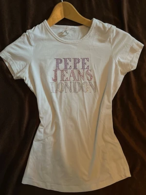 Vit t-shirt från Pepe Jeans London med rhinestones  - Snygg vit t-shirt från Pepe Jeans London med rhinestone text framtill i olika färger. Klassisk rund halsringning och korta ärmar. Perfekt till jeans eller kjol för en trendig look.