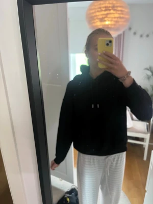 Svart hoodie med huva och ficka - En klassisk svart hoodie med huva och dragsnören. Hoodien har en stor magficka framtill och ribbade muddar vid ärmslut och nederkant. Perfekt för en avslappnad och chill stil. Materialet ser ut att vara mjukt och bekvämt, troligen bomull eller bomullsblandning.