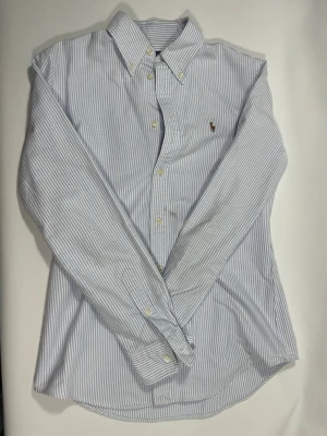 Blårandig skjorta med knappar - Klassisk skjorta med smala blå och vita ränder, button-down krage och långa ärmar. Skjortan har knappar framtill och en liten broderad logga på bröstet. Perfekt för en stilren och avslappnad look. 