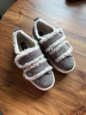 Grå sneakers med fuskpäls från Duffy - Grå sneakers från Duffy med tre breda kardborreband och fluffig vit fuskpäls längs kanterna. Ovandelen är i mockaimitation och sulan är vit. Insidan är fodrad med mjukt material för extra komfort. Perfekt för dig som vill ha något både snyggt och mysigt. Näst intill helt nya. Använt två gånger, därav lite slit på sulan💕 nypris 799kr