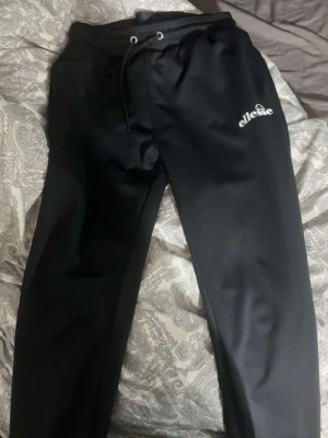 Ellesse byxor - Snygga svarta mjukisbyxor från Ellesse med dragsko i midjan och logga på vänster lår. Byxorna har en avslappnad passform och är perfekta för chill dagar. Tillverkade i mjukt material som känns skönt mot huden.