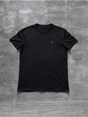 Svart t-shirt med röd logga - Snygg svart t-shirt med rund hals och en liten röd broderad logga på bröstet. Klassisk passform och tillverkad i mjuk bomull som känns skön mot huden. Perfekt till jeans eller shorts för en clean och enkel stil.