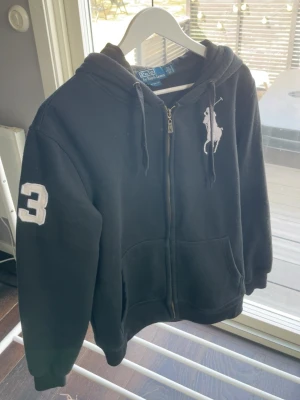 Polo Ralph Lauren hoodie – svart - Svart hoodie med dragkedja från Polo Ralph Lauren. Stor vit logga med polospelare på bröstet och en stor vit broderad siffra 3 på ärmen. Strl L men är som strl M. Säljer då den inte kommer till användning längre.