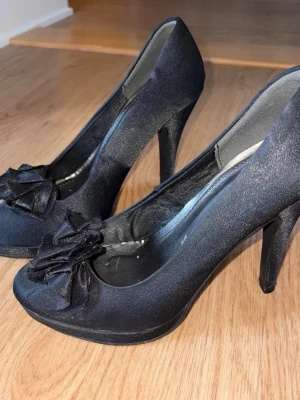 Svarta pumps med rosettdekor - Snygga svarta pumps med hög klack och dekorativa rosetter framtill. Skorna har en klassisk spetsig tå och är tillverkade i ett glansigt tygmaterial som ger en elegant look. Perfekta för att lyfta vilken outfit som helst.