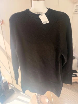 Svart stickad tröja med rund hals - En enkel och stilren svart stickad tröja med rund halsringning och långa ärmar. Tröjan har ribbstickade muddar vid ärmslut och nederkant, vilket ger en klassisk look. Perfekt att matcha med jeans eller kjol för en avslappnad stil.